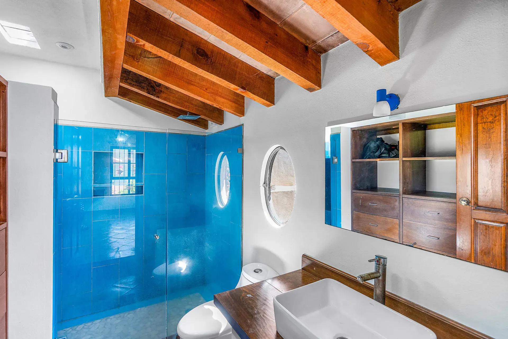 Casa Sirena, bathroom with a bright blue tile shower, an oval window, a vessel sink and a wooden cabinet, San Miguel de Allende