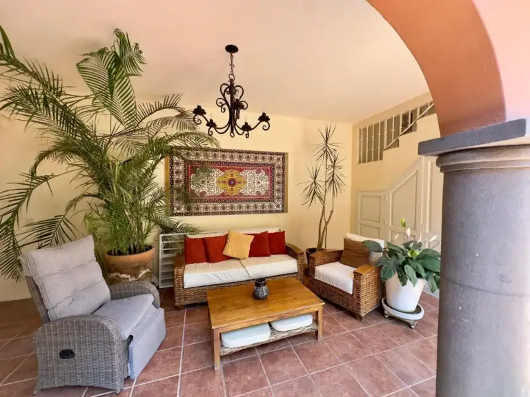Casa Trebol, sitting room with a wicker sofa, armchairs, a coffee table, palms and a woven wall hanging seen through a stone arch, San Miguel de Allende