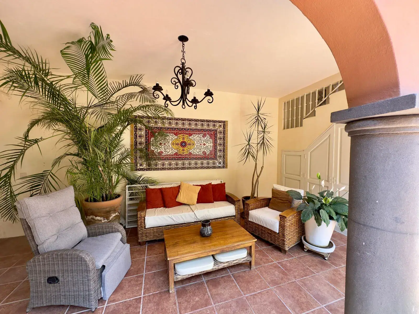 Casa Trebol, sitting room with a wicker sofa, armchairs, a coffee table, palms and a woven wall hanging seen through a stone arch, San Miguel de Allende