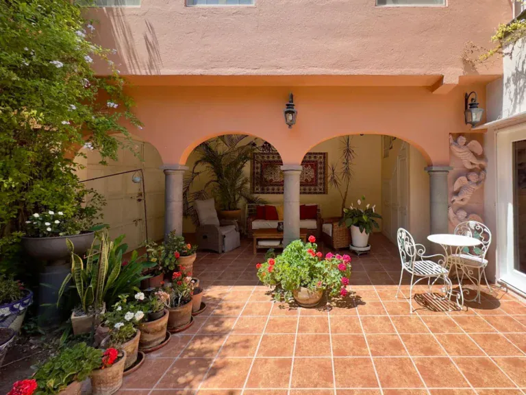 Casa Trebol, covered terrace with two arches, stone columns, a wicker sitting area and potted geraniums on a tile patio, San Miguel de Allende