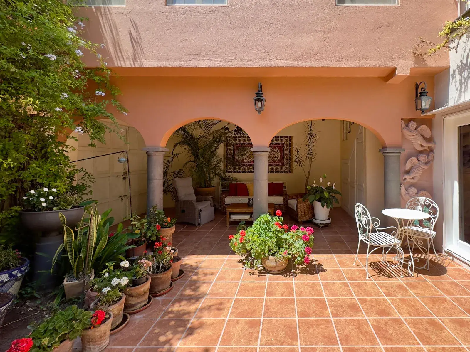 Casa Trebol, covered terrace with two arches, stone columns, a wicker sitting area and potted geraniums on a tile patio, San Miguel de Allende