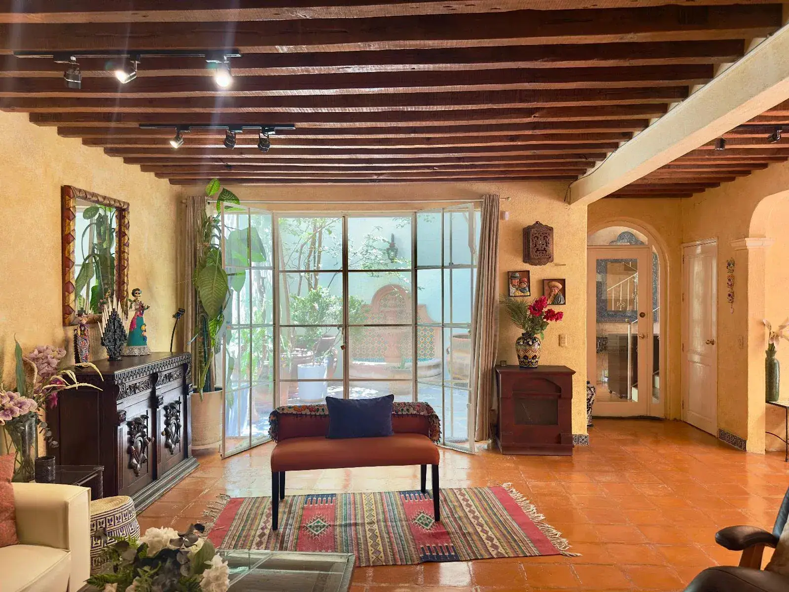 Casa Trebol, living room with a dark wood timber ceiling, a steel-framed glass wall, a carved wood chest and a red rug, San Miguel de Allende