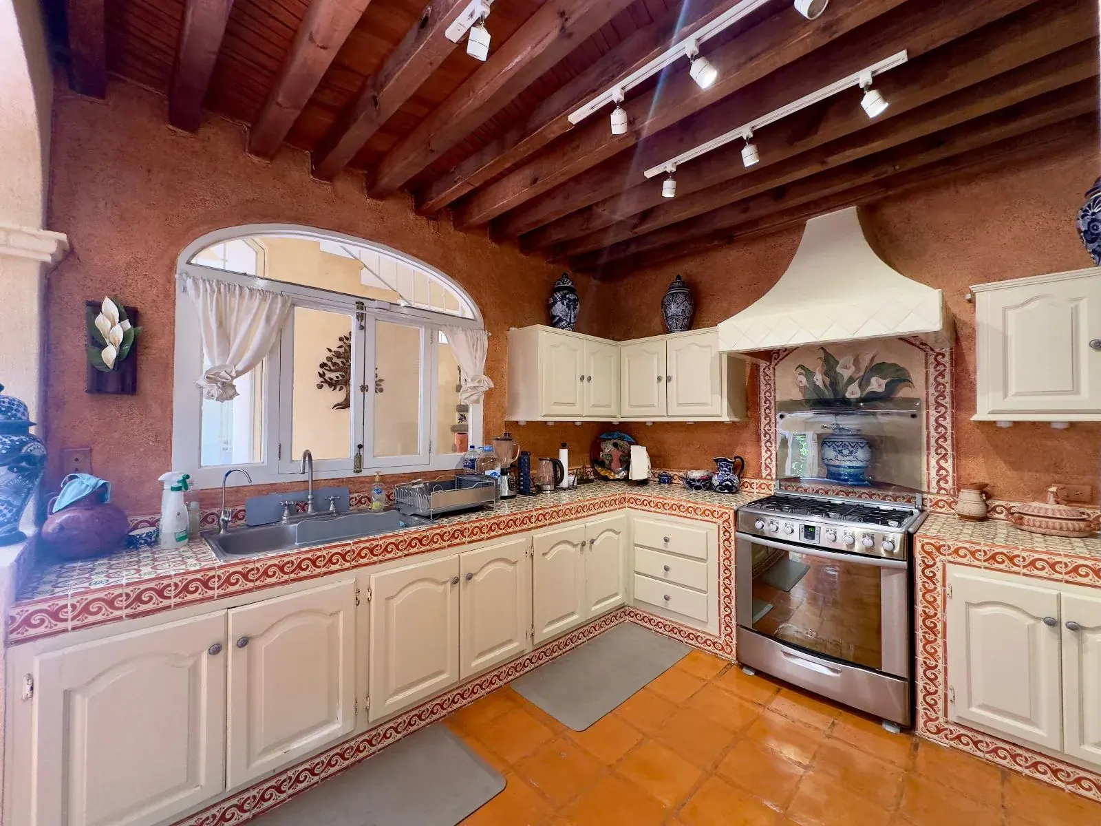 Casa Trebol, kitchen with cream cabinets, hand-painted tile counters, a stainless gas range, a white hood and a timber ceiling, San Miguel de Allende