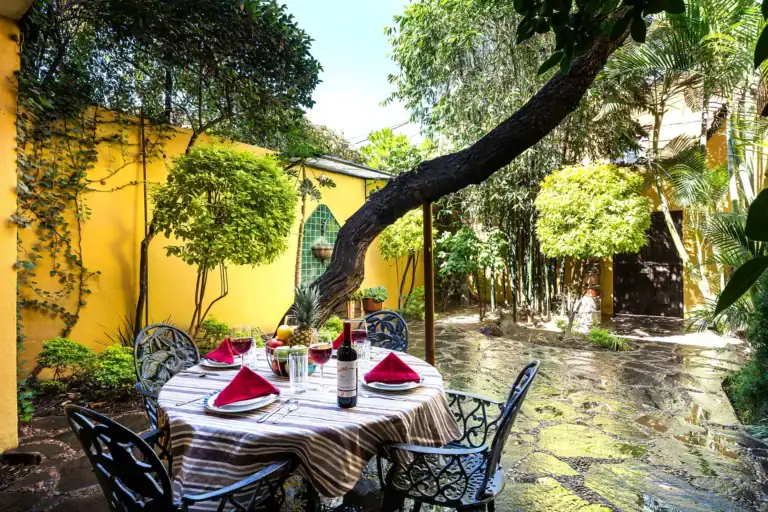 Casa Victoria, courtyard garden with a round table under a leaning tree and a yellow wall, San Miguel de Allende