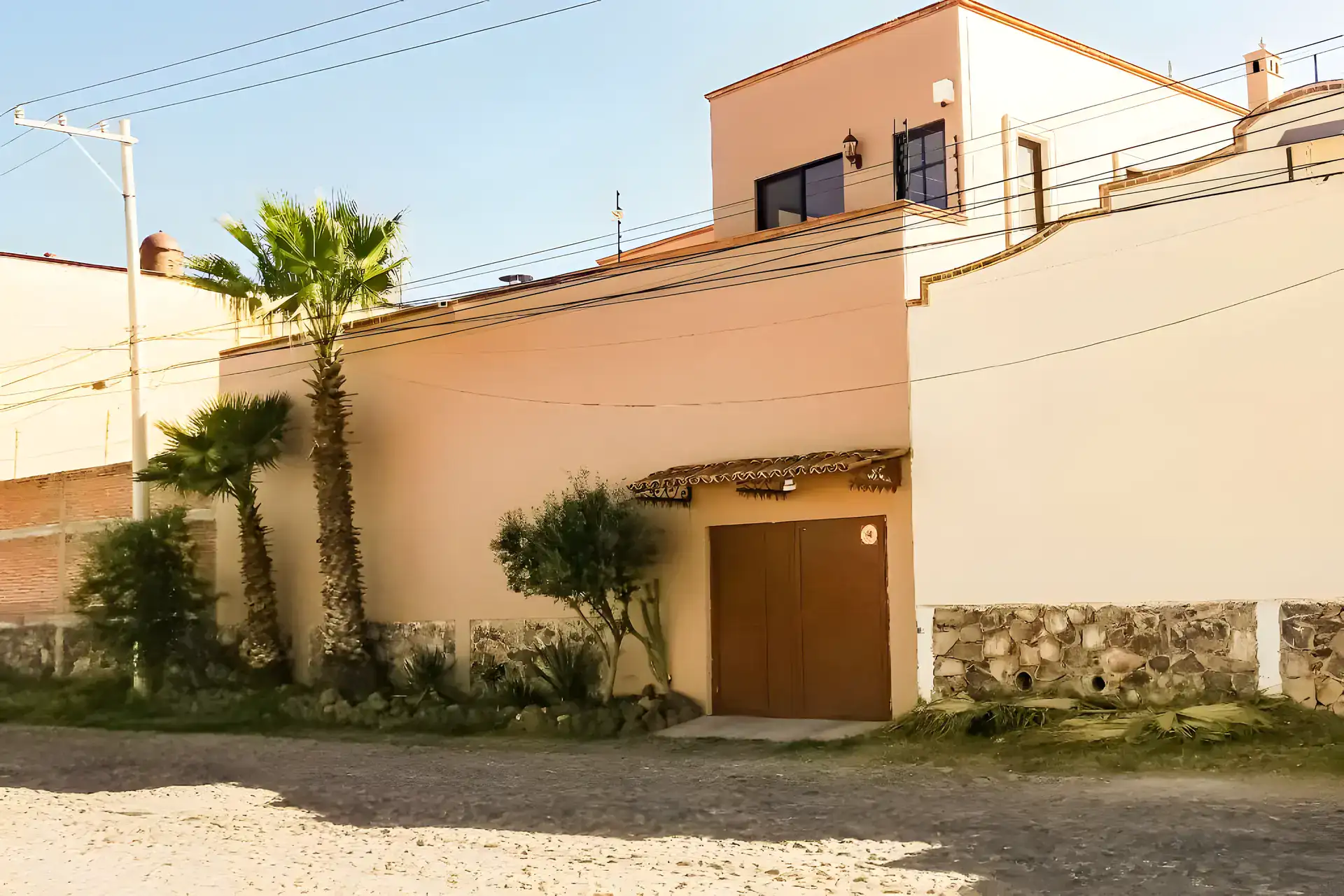 Casa Winston, rear of the house with a palm tree, a wall with stonework and a garage door on a gravel lane, San Miguel de Allende