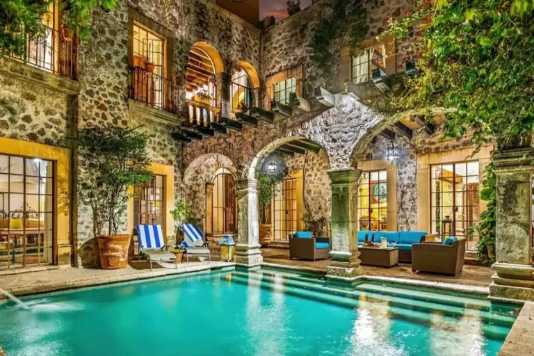 Stone colonial-style home with an arched colonnade and an illuminated swimming pool in a courtyard at dusk