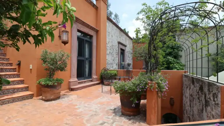 Sunny terracotta courtyard with potted plants, tiled stairs, and a metal arched trellis beside a stone wall on a sunny day, San Miguel de Allende