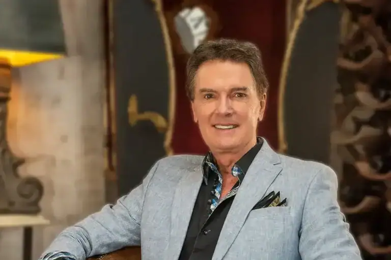 Greg Gunter seated in a brown leather armchair in a light gray blazer, dark shirt and white trousers