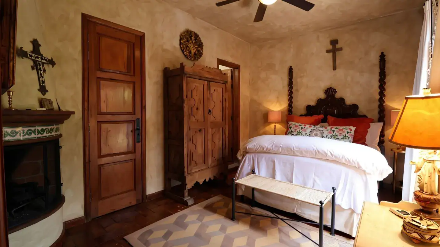 Cozy rustic bedroom with a carved wooden four-poster bed, tall armoire, beige walls, and a fireplace with a green-tiled mantel, San Miguel de Allende
