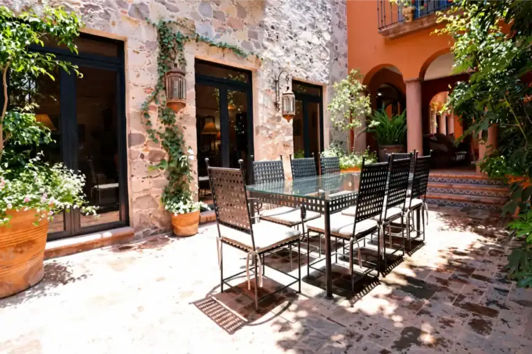 Outdoor courtyard dining area with a glass-topped table and six black metal chairs, sunlit stone walls, and potted plants nearby, San Miguel de Allende