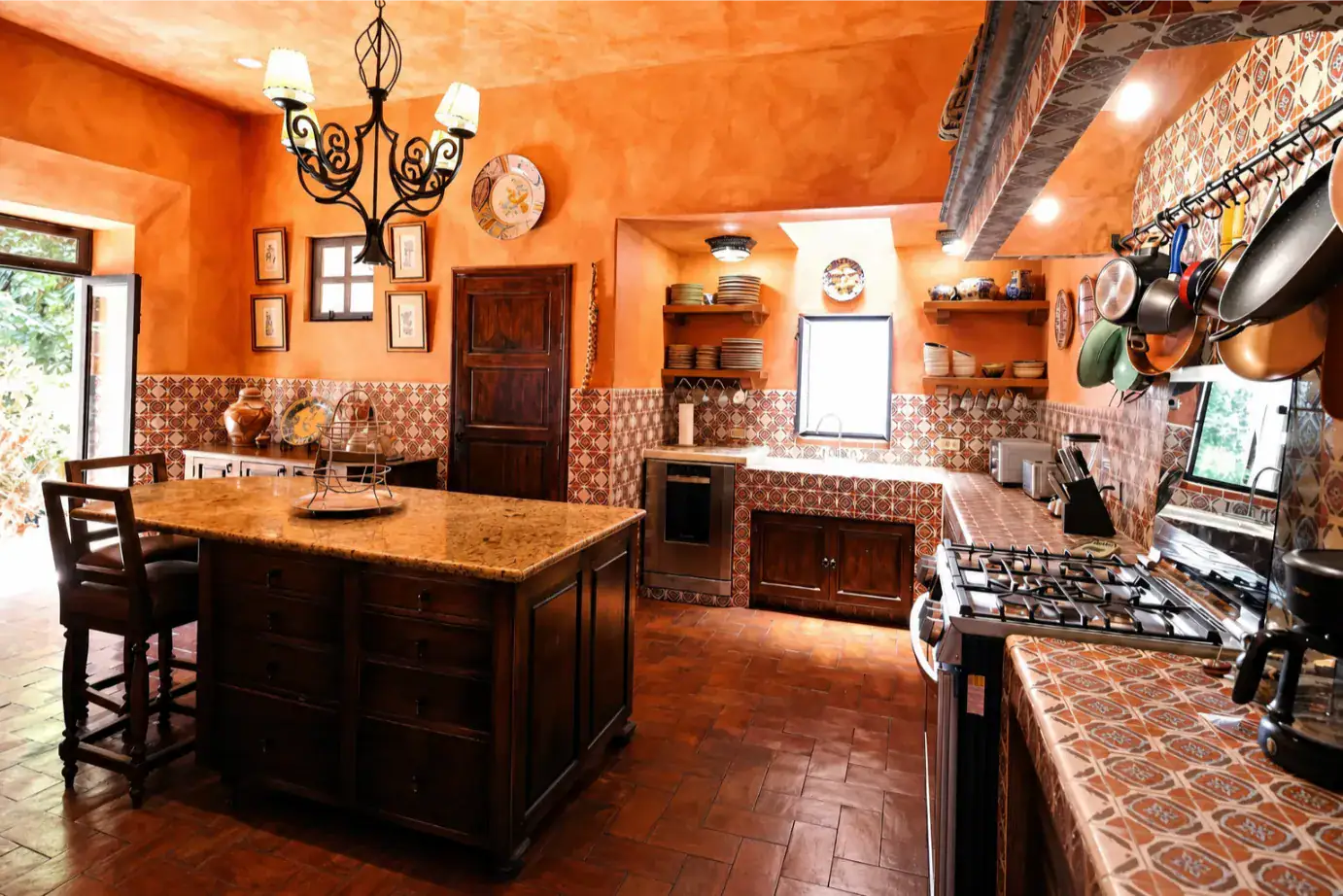 Warm orange-toned kitchen with a large wooden island, tiled countertops, and wall shelves holding dishes and decor, San Miguel de Allende
