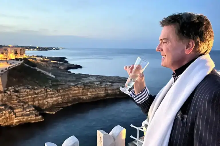 Greg Gunter raising a glass at sunset on a terrace overlooking a rocky coastline