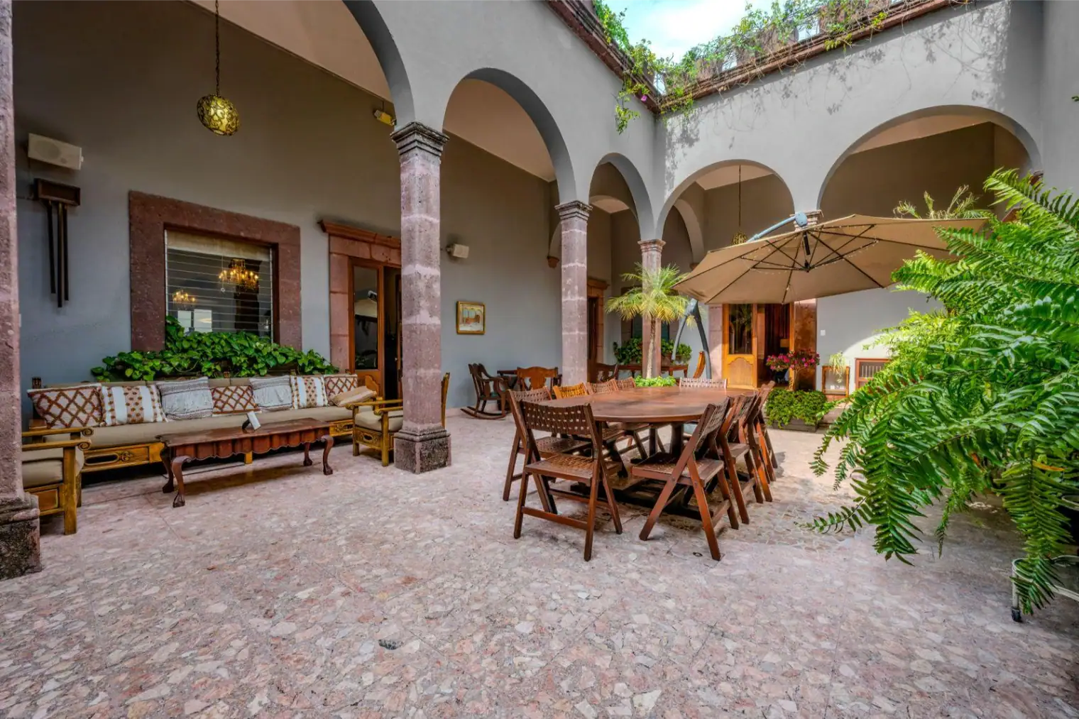 Courtyard with arched colonnade, wooden dining table and chairs under a large umbrella, and cushioned seating along the wall with potted plants nearby, San Miguel de Allende