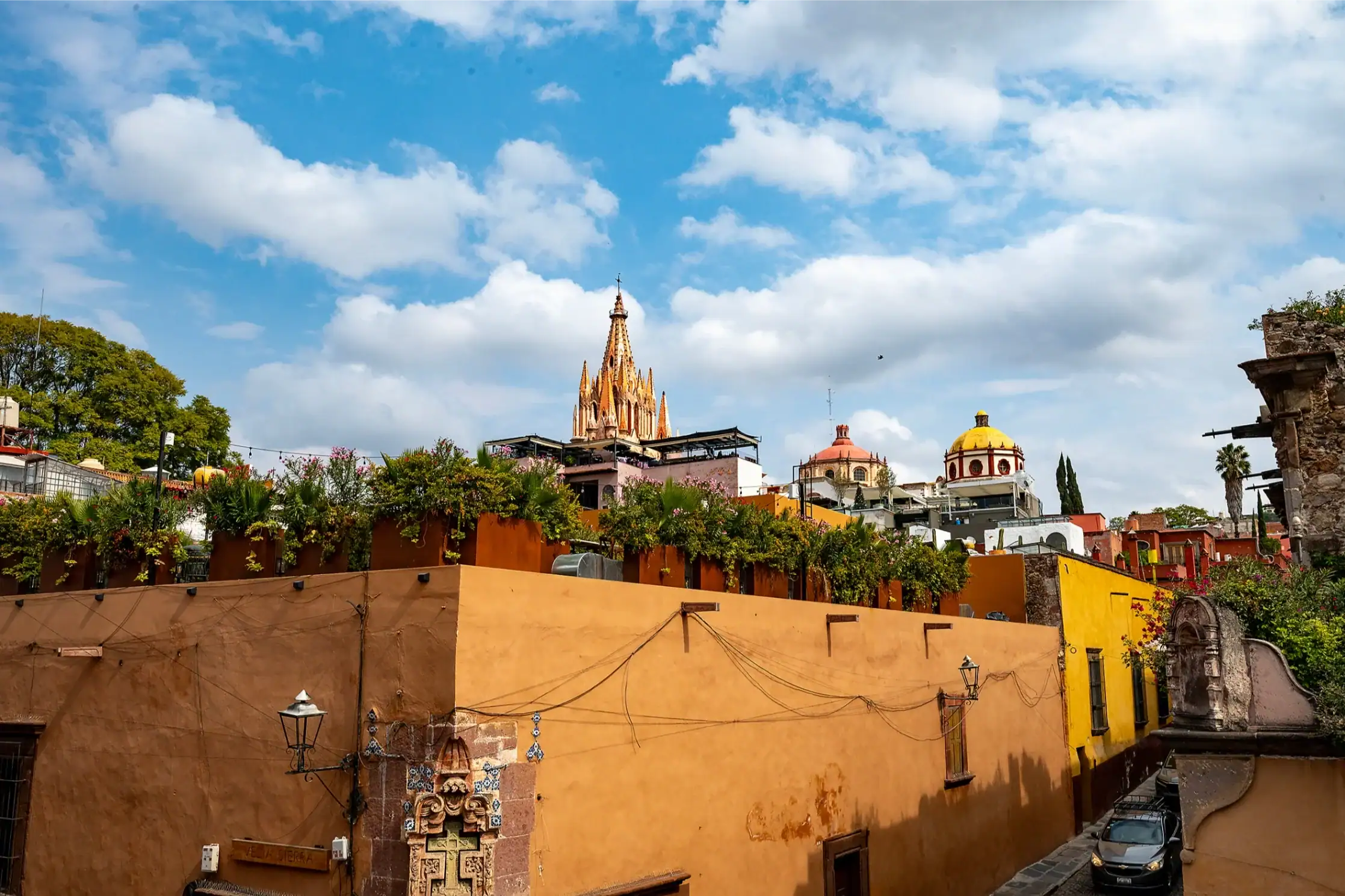 Vivid street scene with warm orange walls, greenery, and a tall orange cathedral spire rising against a blue, partly cloudy sky, San Miguel de Allende
