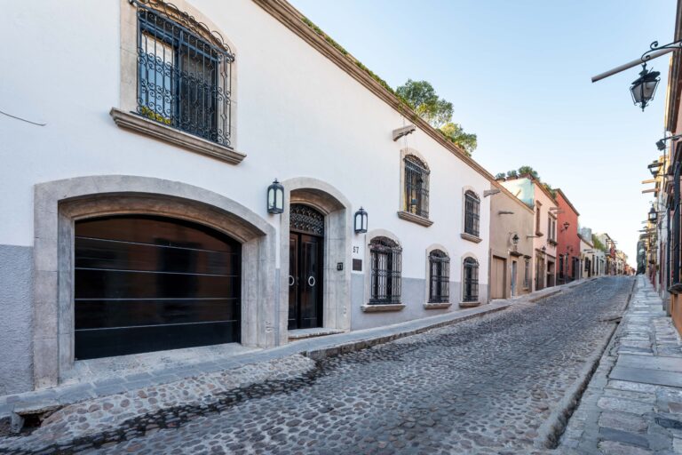 Narrow cobblestone street lined with white stucco buildings, decorative wrought-iron window bars, and arched doorways on a sunny day, San Miguel de Allende