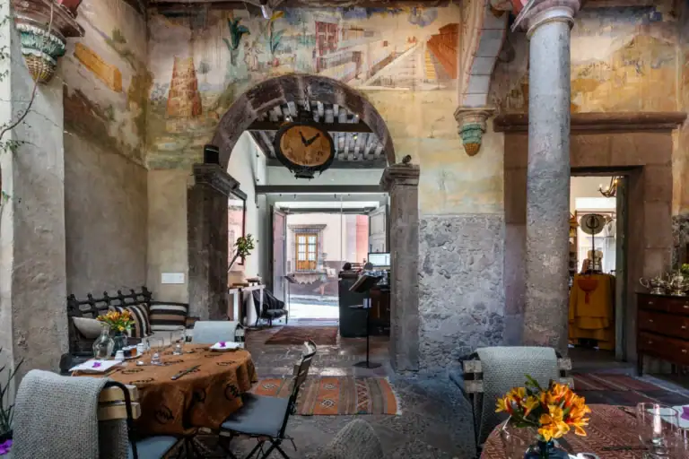 Casa No Name, courtyard dining room with a painted fresco wall, a stone arch with a hanging clock, set round tables with flowers and the street entrance beyond, San Miguel de Allende