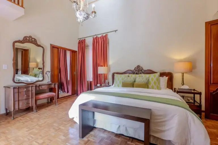 Elegant bedroom with a carved wooden headboard, matching dresser and mirror, and burgundy drapes under a crystal chandelier, San Miguel de Allende