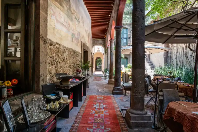 Covered stone alley cafe with red patterned rug, wooden beams, and outdoor seating under umbrellas, San Miguel de Allende