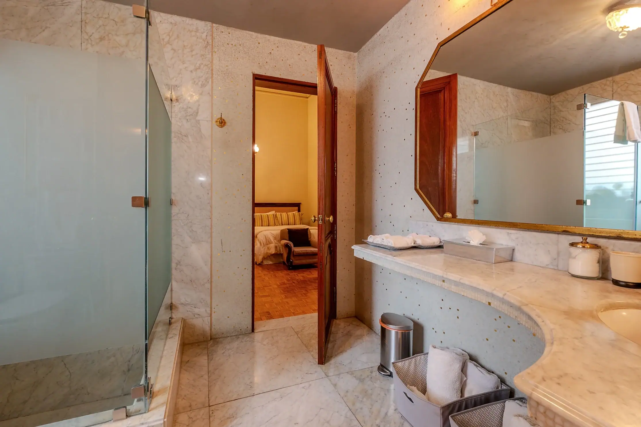 Luxurious marble bathroom with a glass shower on the left, a wide marble counter, and an open wooden doorway into a bedroom with a bed visible through the frame, San Miguel de Allende