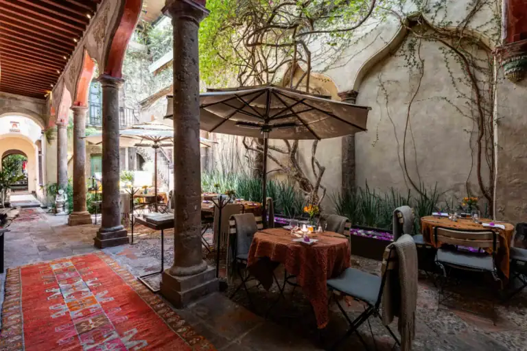 Outdoor restaurant patio with stone columns, archways, and tables set under large umbrellas in a sunlit courtyard, San Miguel de Allende