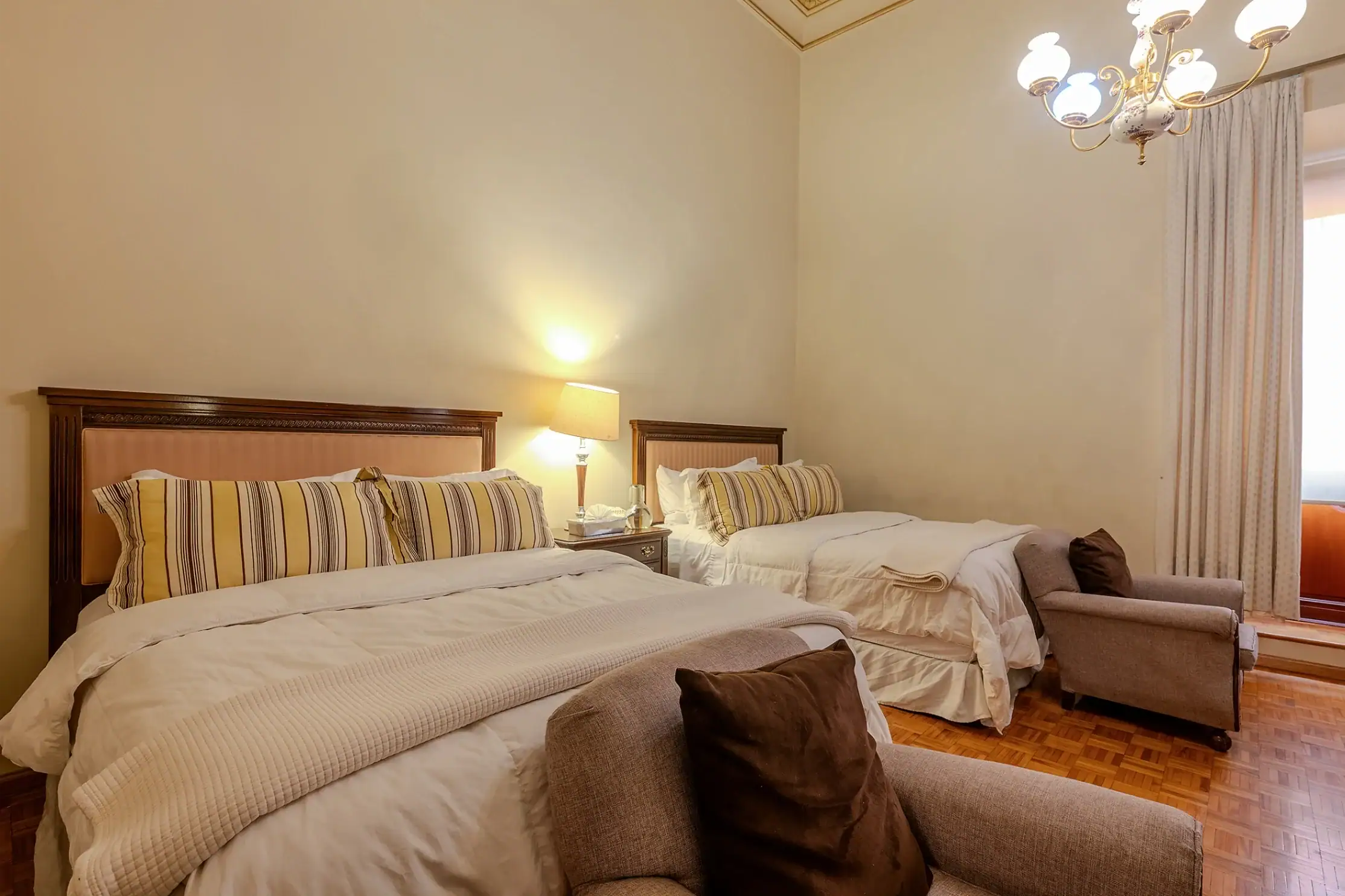 Hotel bedroom with two twin beds, striped pillows, and matching headboards; bedside table lamp between them; chandelier overhead and beige walls with curtains, San Miguel de Allende