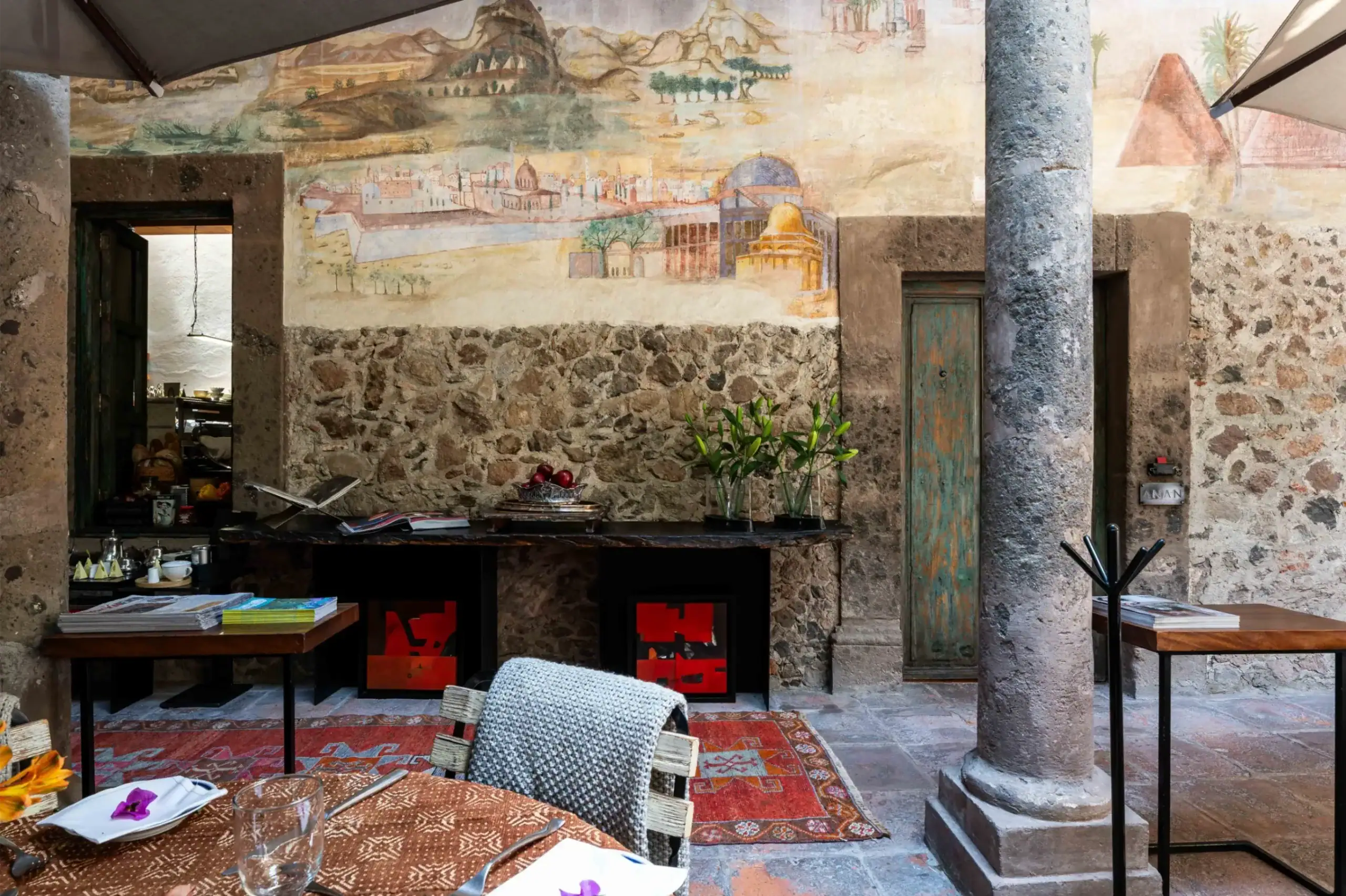 Outdoor dining area with rustic stone walls, a mural, and tall columns; tables set with dishes, glasses, and napkins, San Miguel de Allende