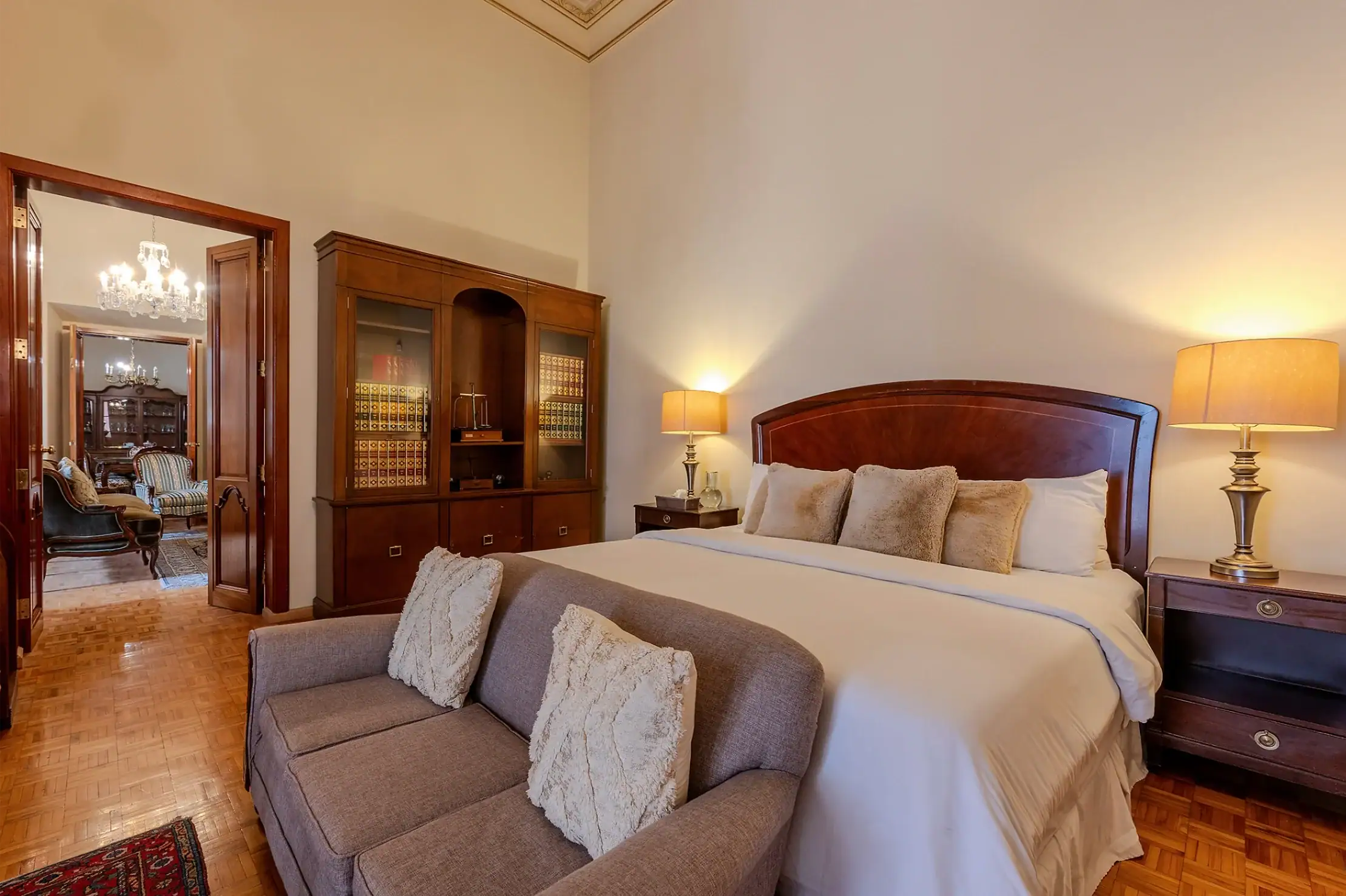 Elegant hotel bedroom with a large wooden headboard, neutral bedding, and plush pillows, flanked by matching lamps; a wooden cabinet fills the wall to the left, and a doorway reveals a chandeliered seating area in the distance, San Miguel de Allende