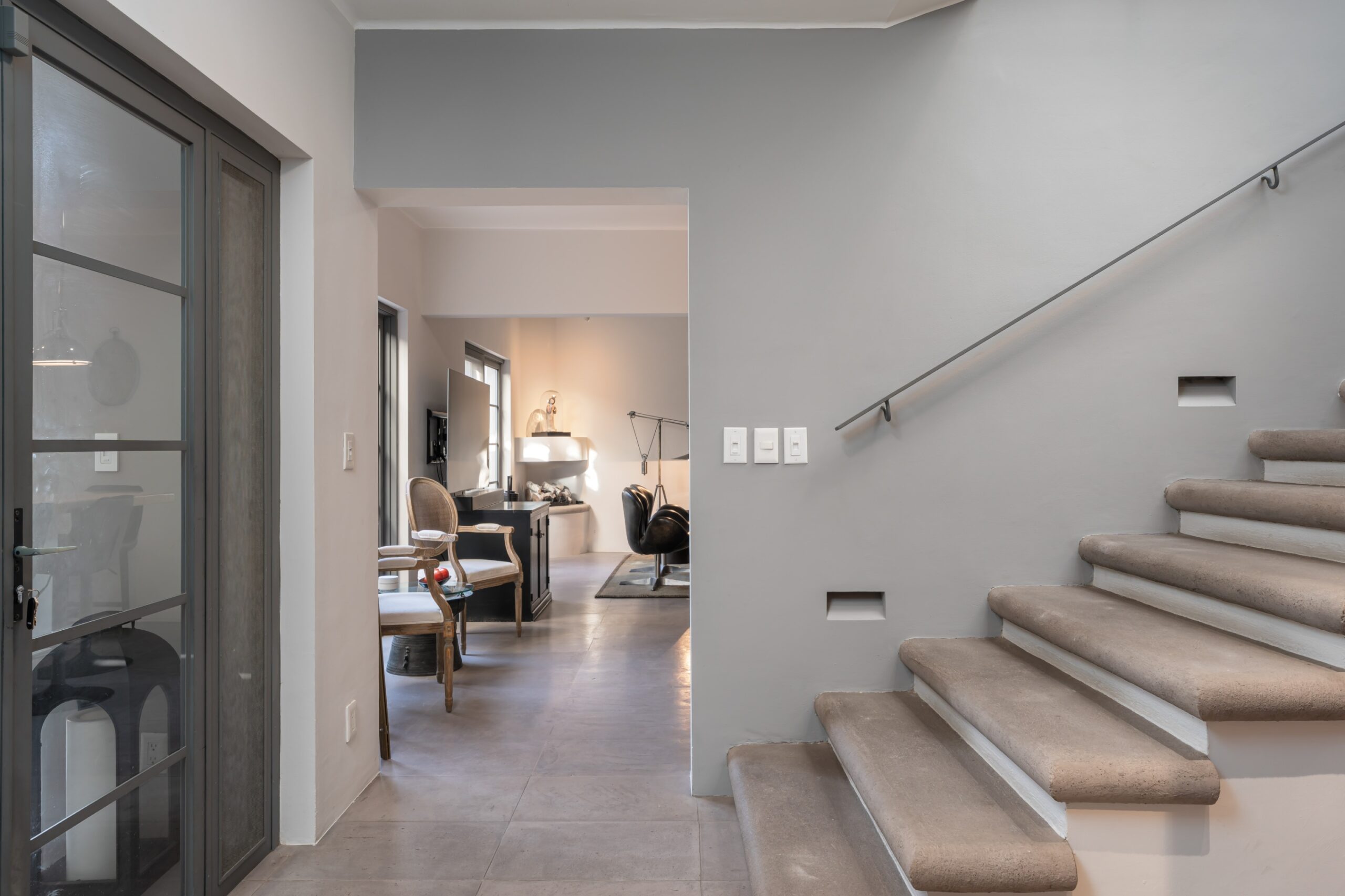 Modern interior hallway with a gray staircase on the right and a glass-pane door on the left, opening to a living area. It shows chairs and a table in the background, San Miguel de Allende