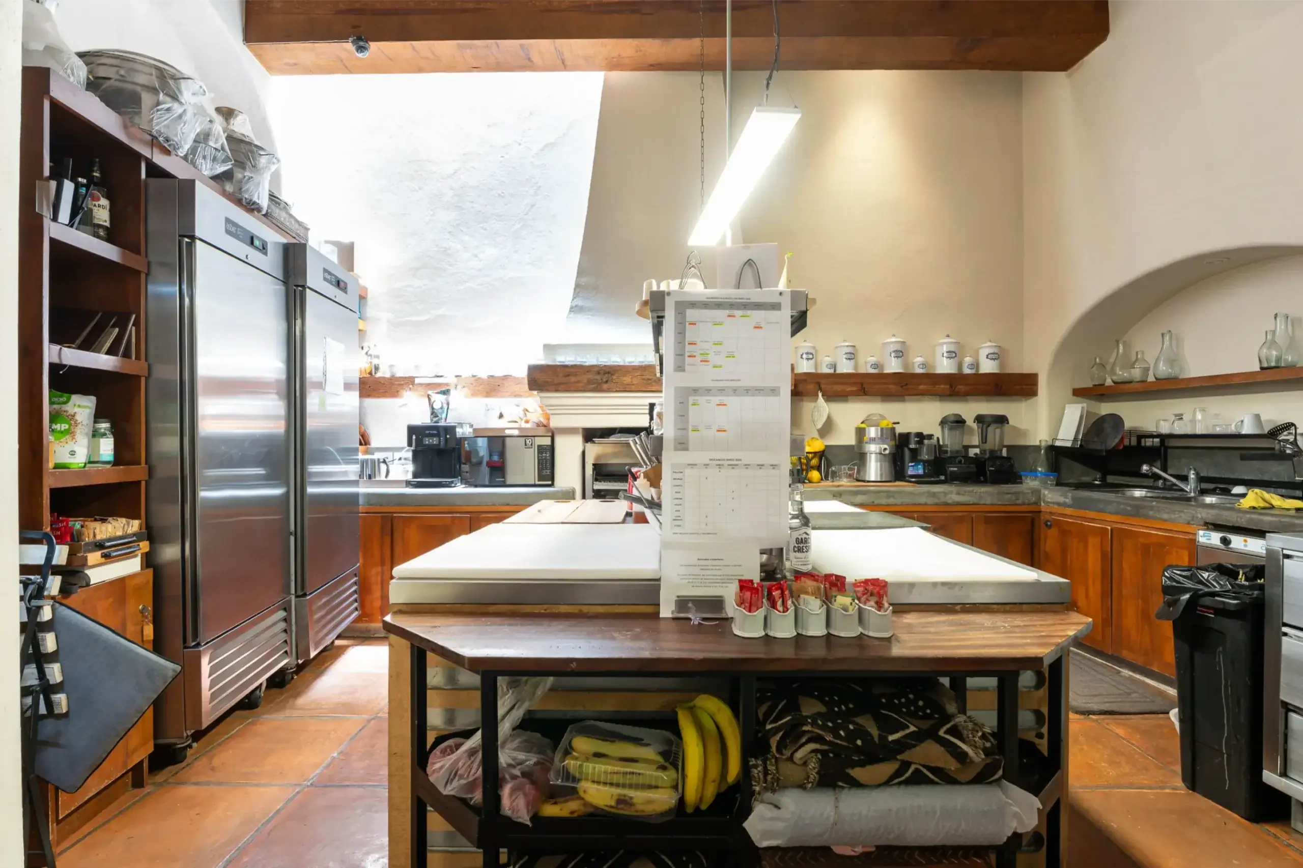 Commercial kitchen scene with stainless steel refrigerators on the left, a central prep table, and shelves with jars and bottles in the background, San Miguel de Allende