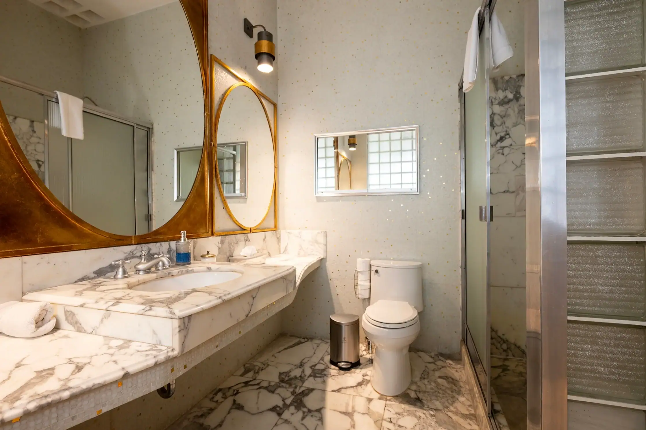Marble bathroom with a double sink vanity, gold oval mirrors, wall sconce, and a glass-enclosed shower, San Miguel de Allende