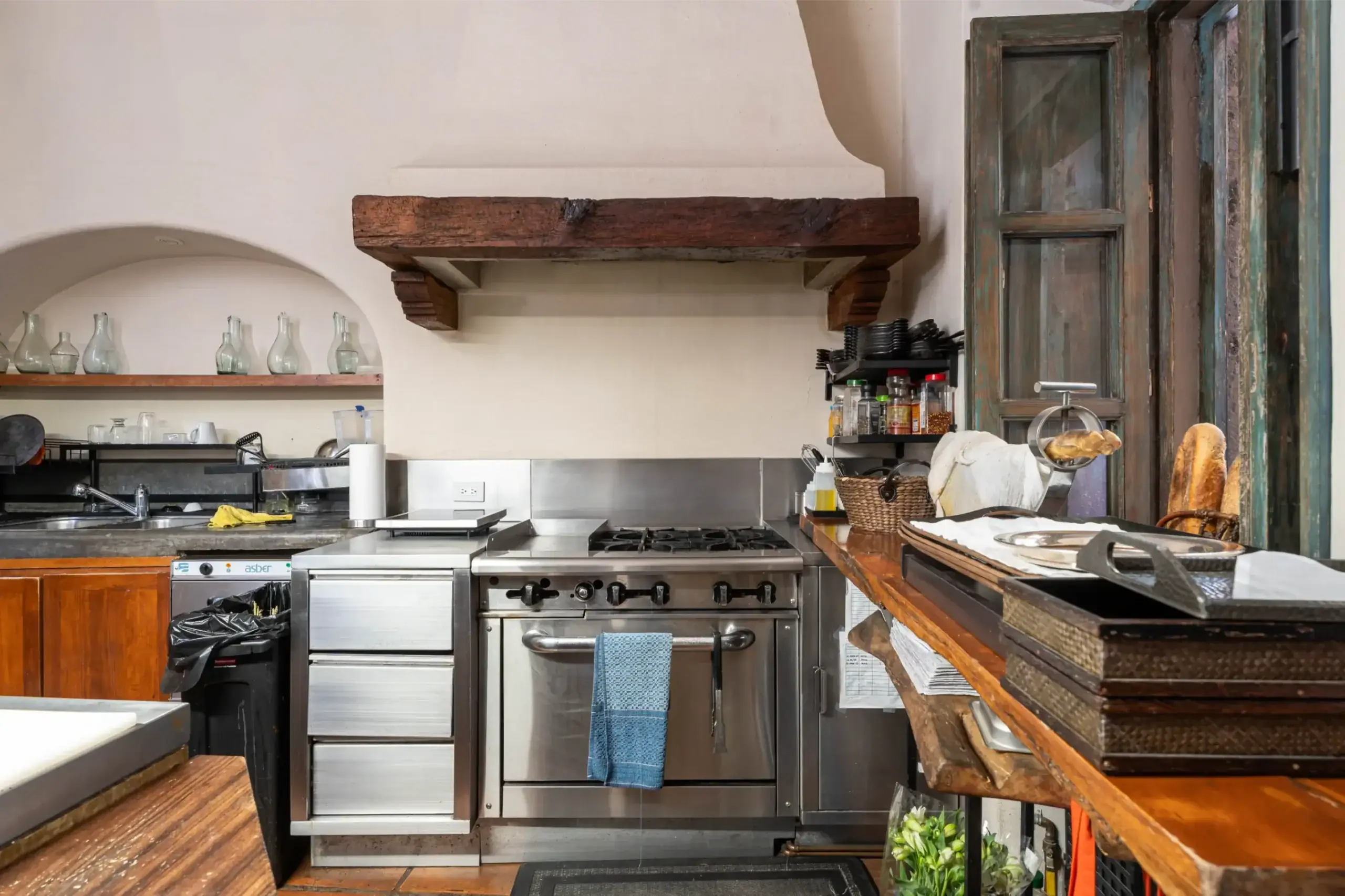 Rustic commercial kitchen with a large stainless steel stove, a heavy wooden beam shelf above, and glass bottles on the left shelf while bread sits on the right counter, San Miguel de Allende