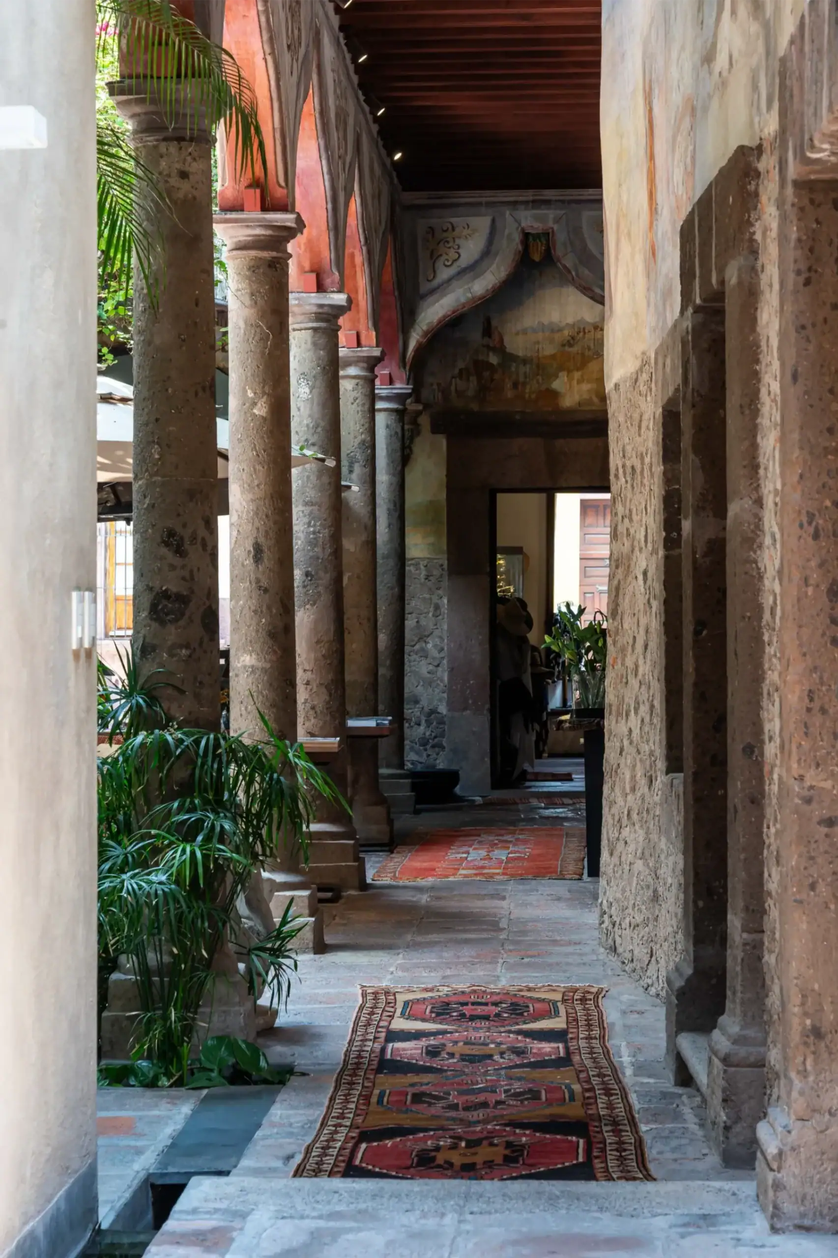 Stone-columned walkway in a historic courtyard with red arches, murals, and patterned floor rugs, San Miguel de Allende
