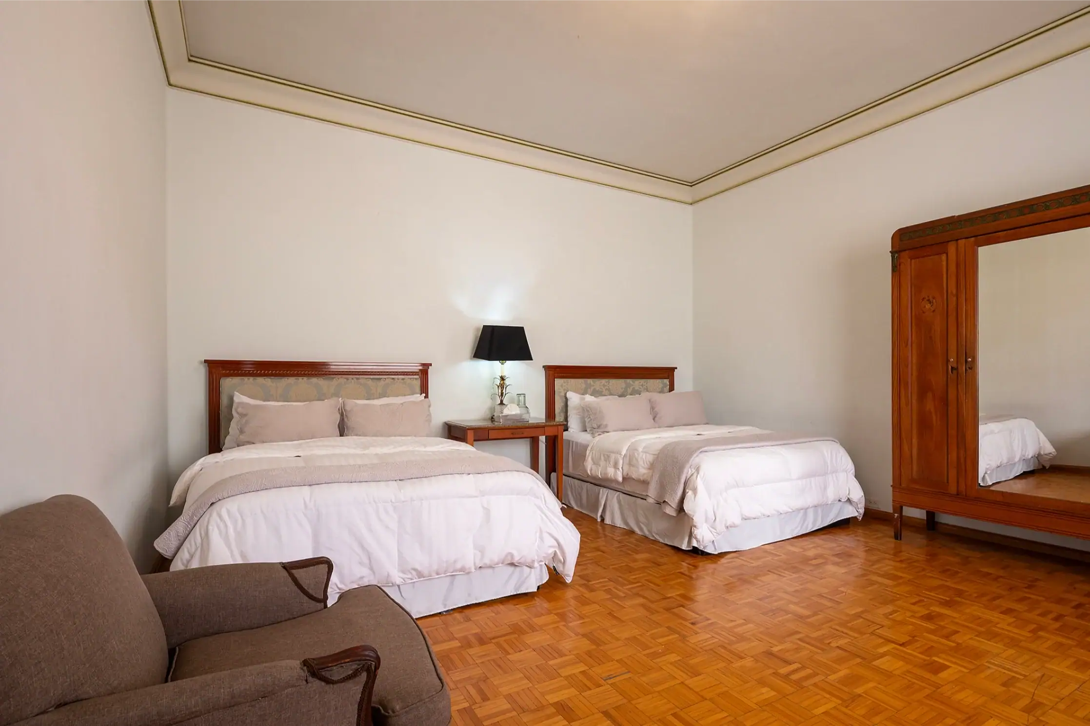 Hotel bedroom with two twin beds, nightstands, lamp, and a wooden armoire against pale walls, San Miguel de Allende