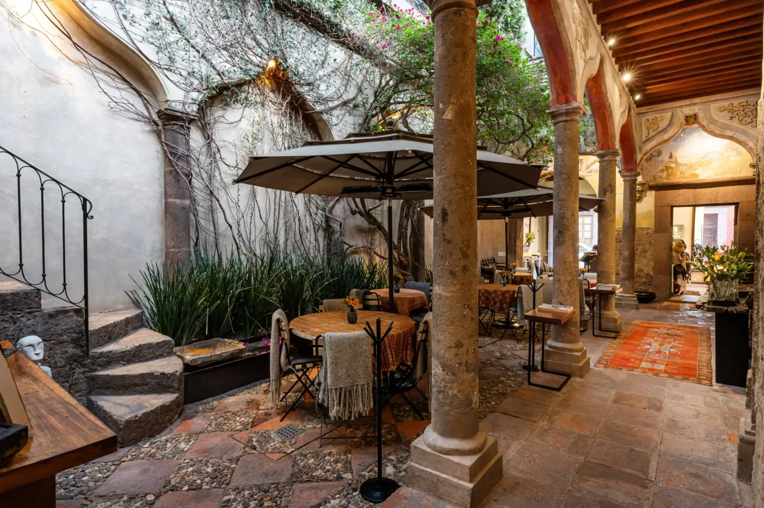 Open-air restaurant courtyard with stone columns, arches, and umbrellas over tables served in a sunlit setting and potted plants nearby, San Miguel de Allende