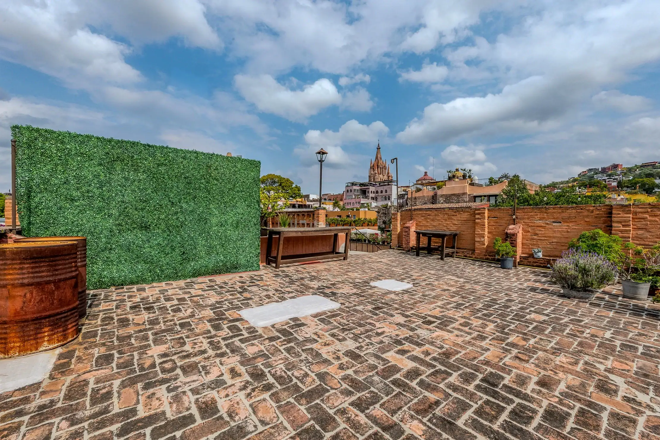 Rooftop terrace with a tall green hedge wall on the left, brick-paved ground, rustic barrels, and wooden seating, skyline with a church spire in the distance under a blue, cloudy sky, San Miguel de Allende