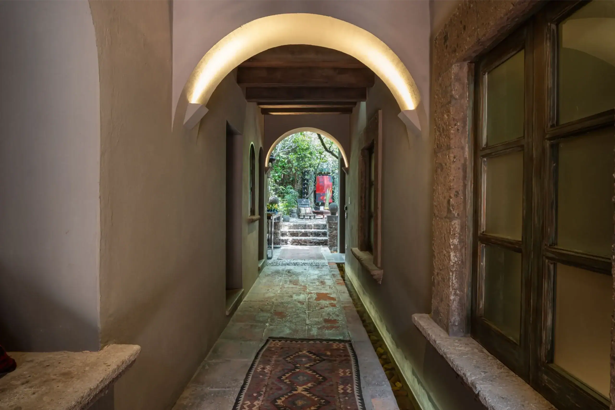 Narrow stone corridor with softly lit arches leading to a sunlit garden courtyard at the end, San Miguel de Allende