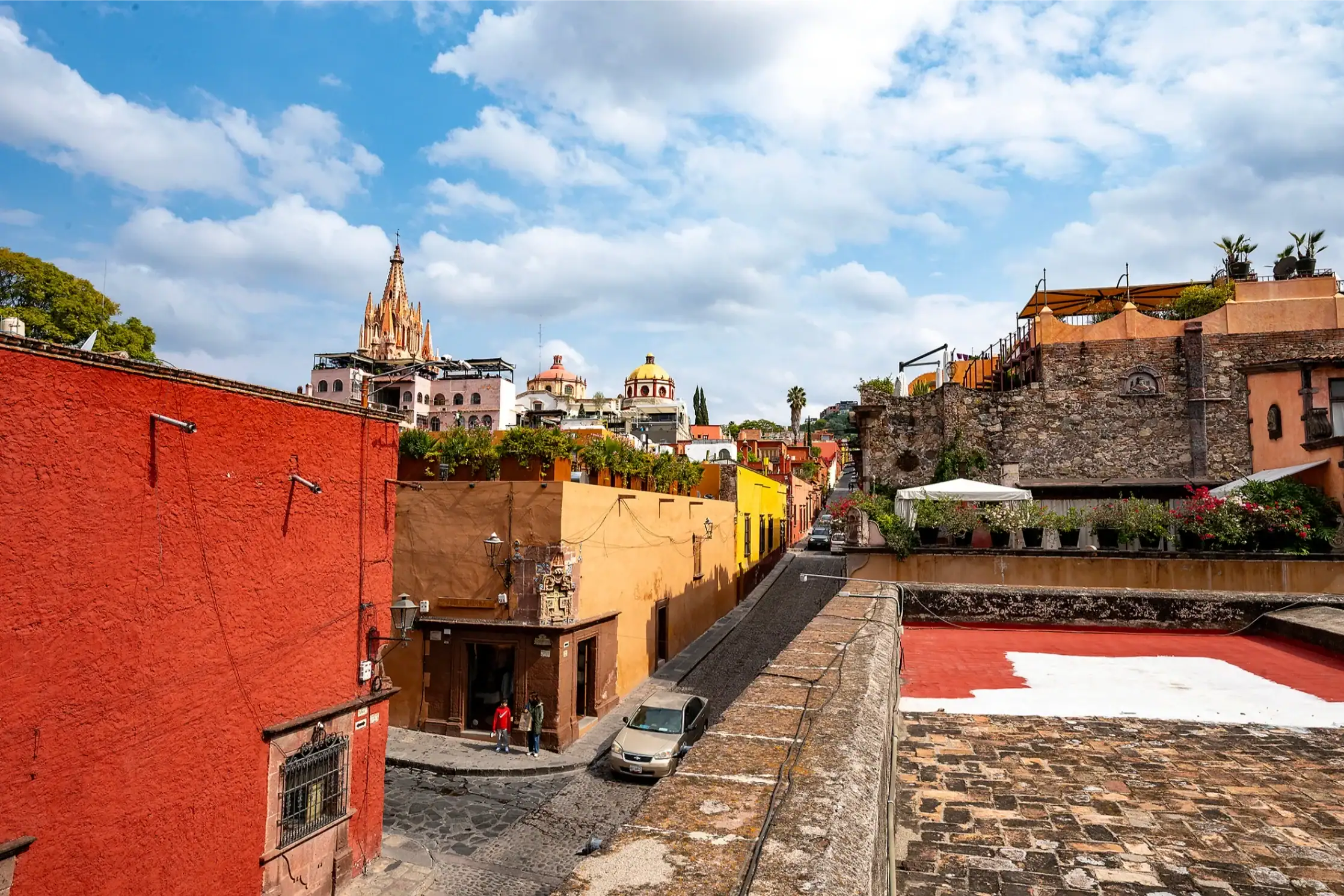 Colorful Mexican street scene with red and orange buildings, a cobblestone road, and a distant church spire under a bright blue sky, San Miguel de Allende