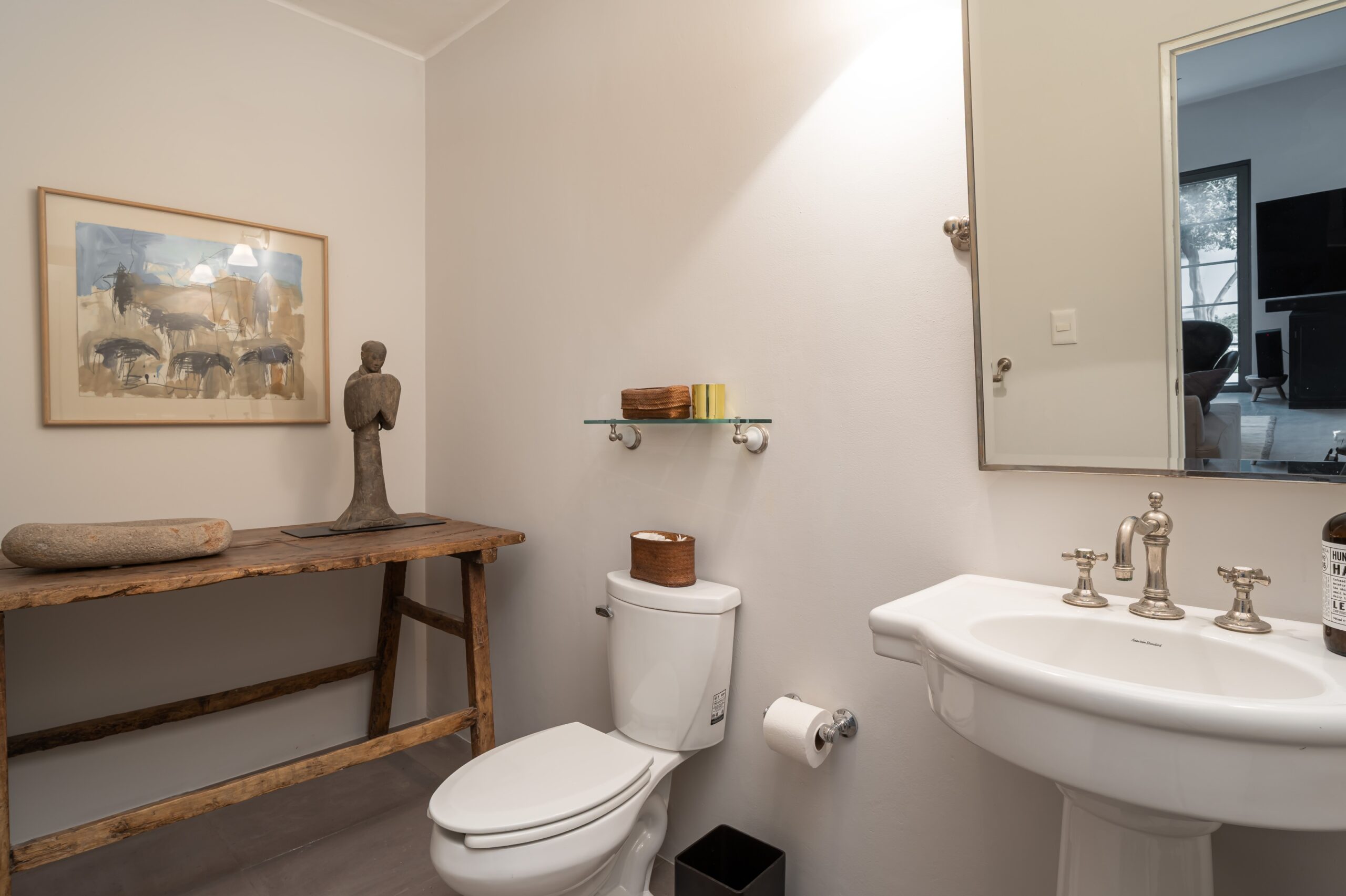 Rustic bathroom corner with a wooden bench, a stone dish, a small abstract sculpture, and a framed abstract painting above the bench, San Miguel de Allende