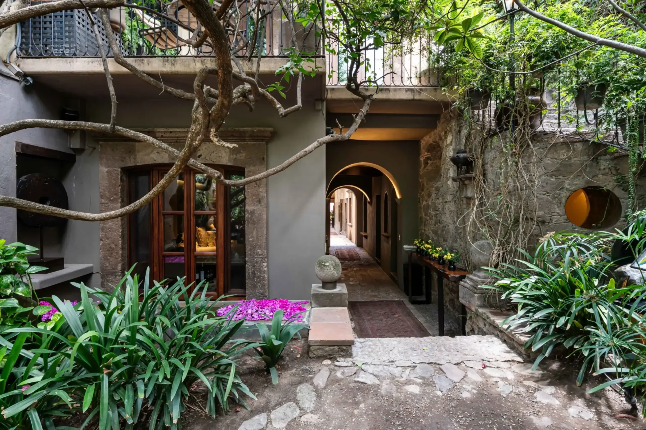Courtyard with stone walls, arched doorways, and lush green plants spilling over branches and vines onto a balcony above, San Miguel de Allende