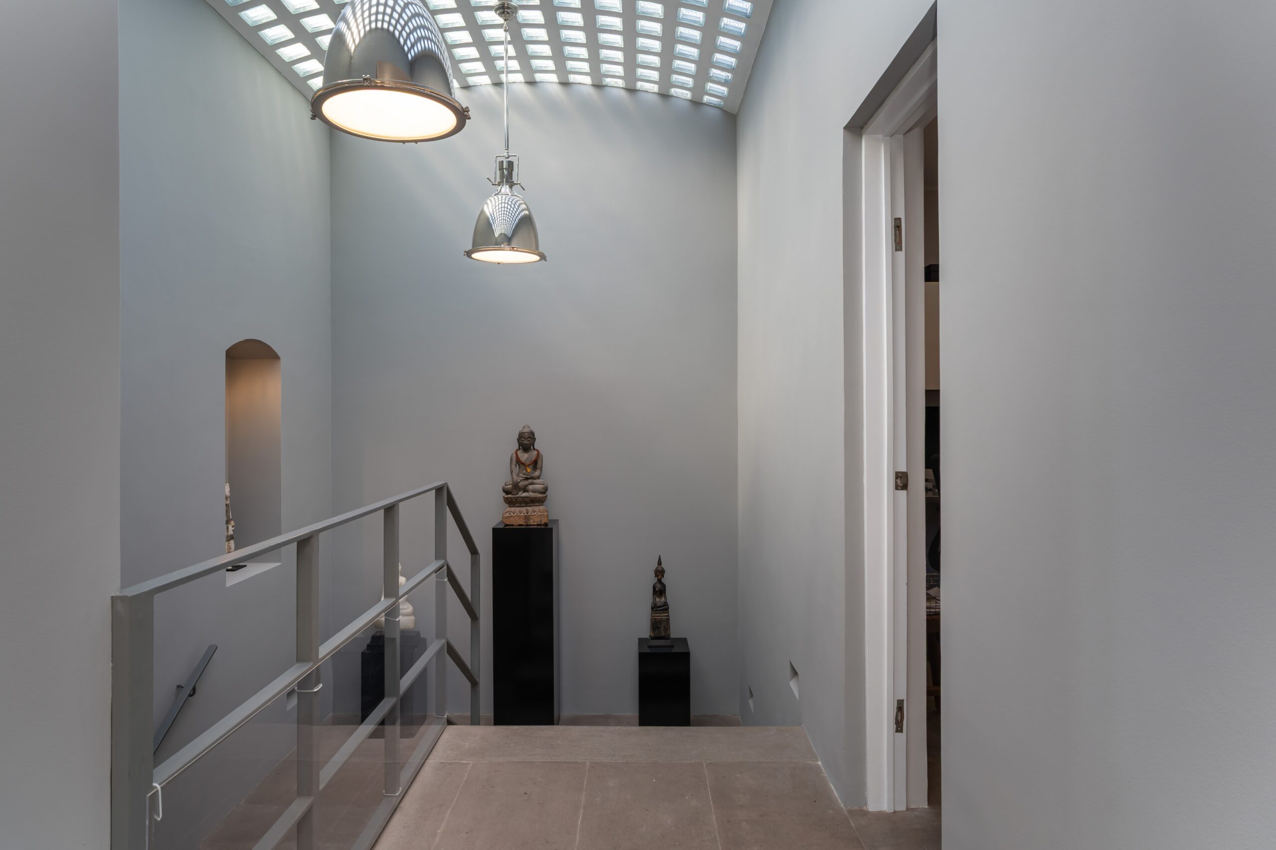 Interior view of a minimalist gallery with statues on black pedestals, glass railing, and two large metallic pendant lights in a pale gray space under a skylight grid ceiling, San Miguel de Allende
