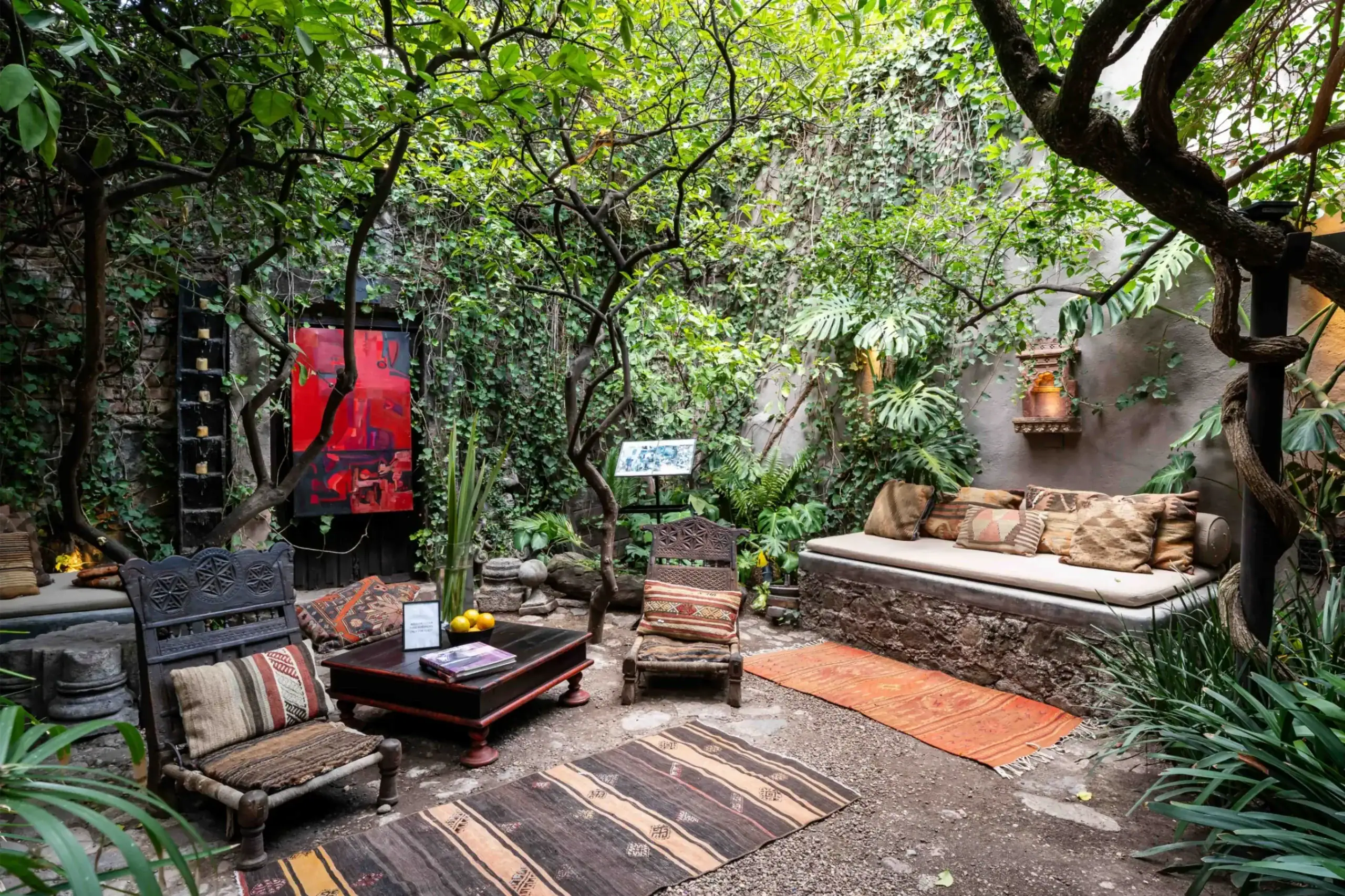 Cozy outdoor garden seating area with patterned rugs, cushions, and low wooden tables amid trees and ivy-covered walls, San Miguel de Allende