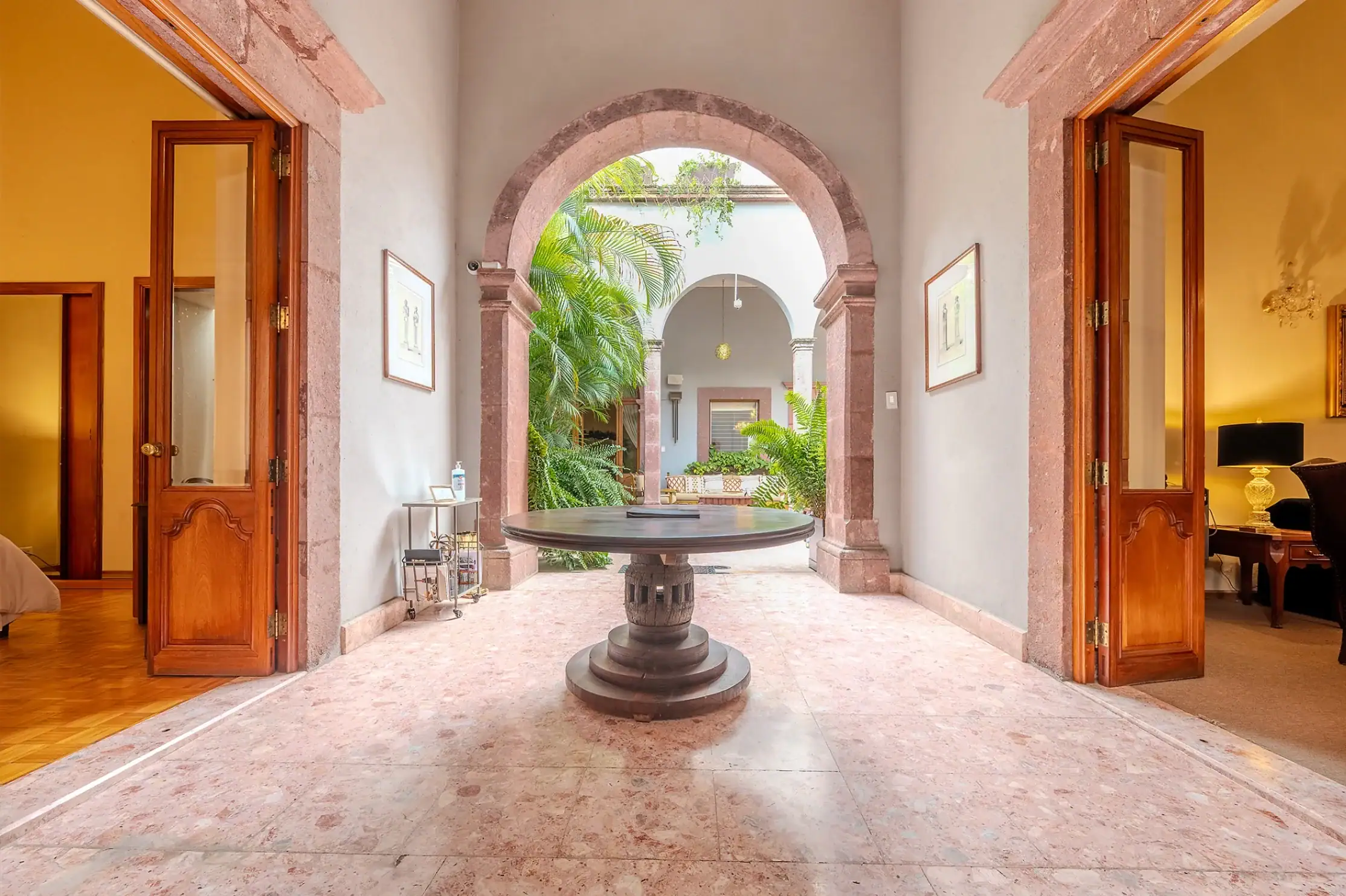 Stone-arched passage opening to a courtyard with tropical plants, a round pedestal table in the foreground, and rooms with wooden doors on both sides, San Miguel de Allende