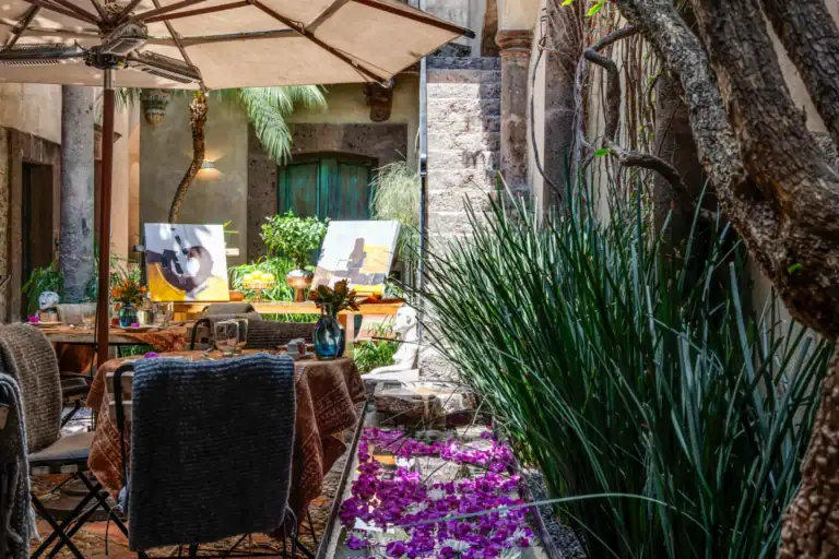 Outdoor courtyard with art easels, tables set for a meal, and purple flower petals along a narrow water feature, San Miguel de Allende