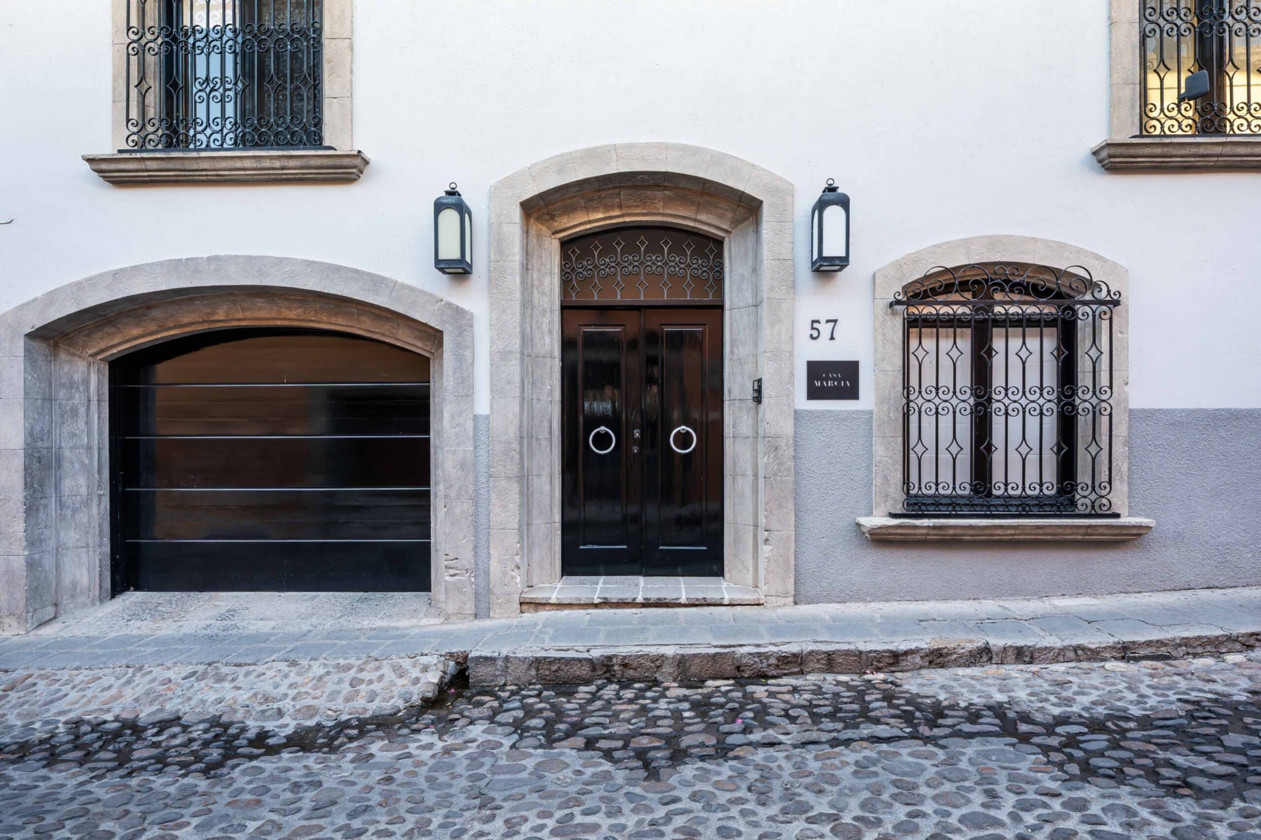 Facade of a pale building with a stone-arched entrance, dark wooden doors, iron grille above, and a cobblestone street in front, San Miguel de Allende