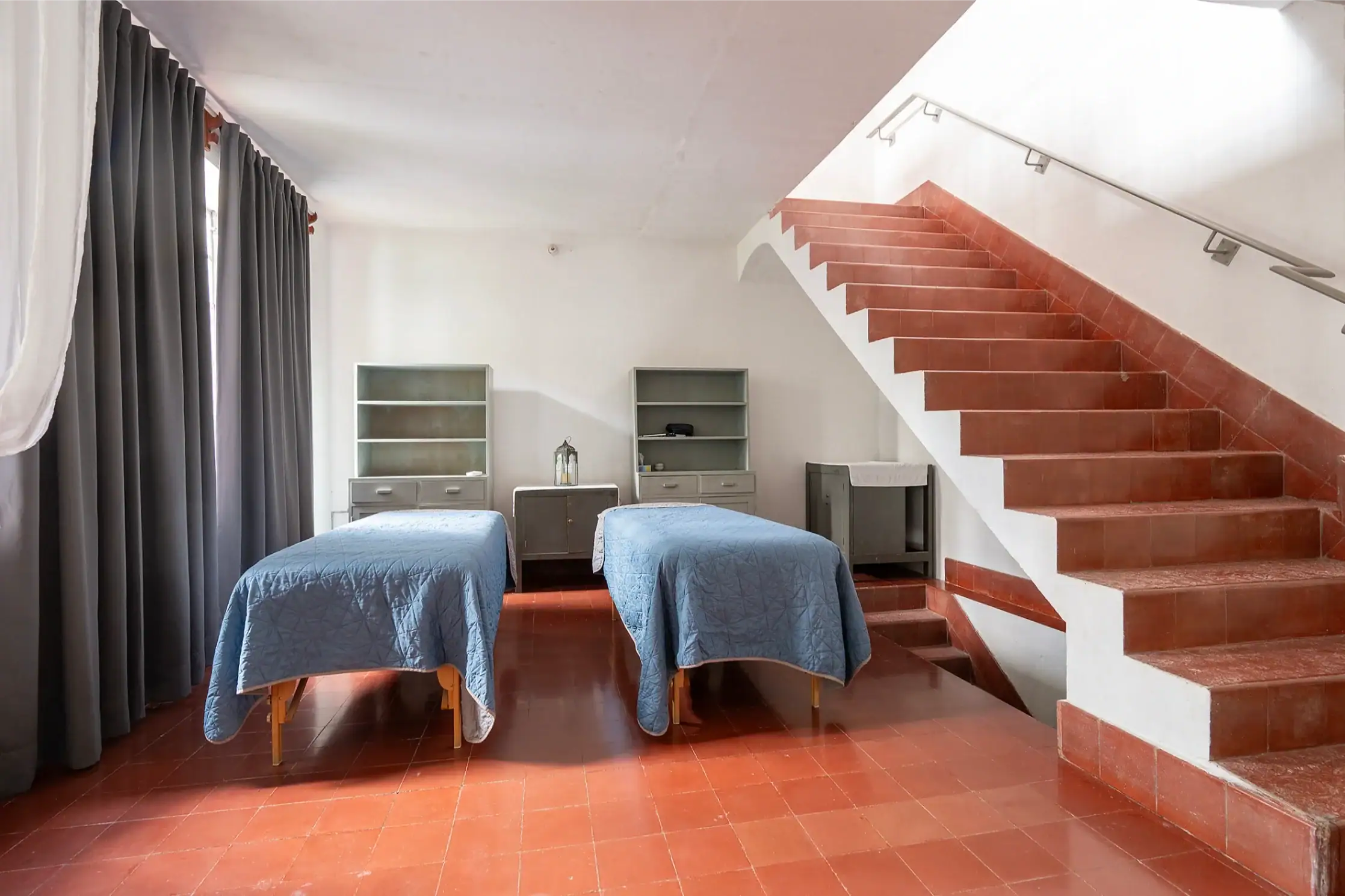 Twin beds with blue quilts sit side by side in a bright room, a red-tiled staircase rises along the right wall, with gray shelving in the background, San Miguel de Allende