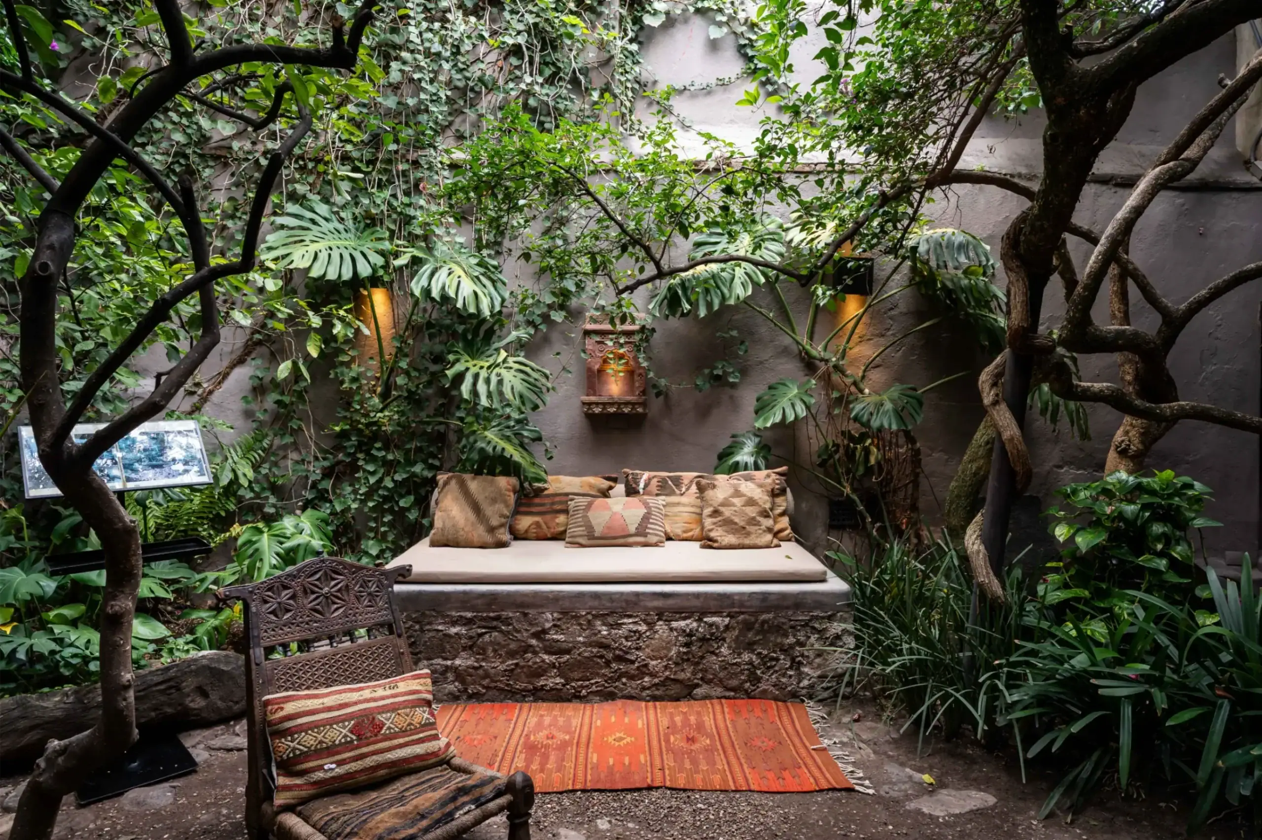 Outdoor garden seating area with a long cushioned daybed and patterned throw pillows, surrounded by lush green tropical plants and vines against a gray wall, San Miguel de Allende