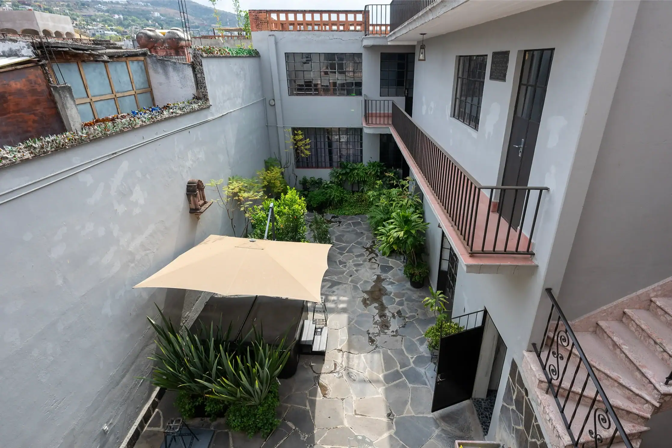 Outdoor courtyard of a multi-story residential building with gray walls, stone-paved ground, potted plants along the walls, and a beige patio umbrella over a small seating area, San Miguel de Allende