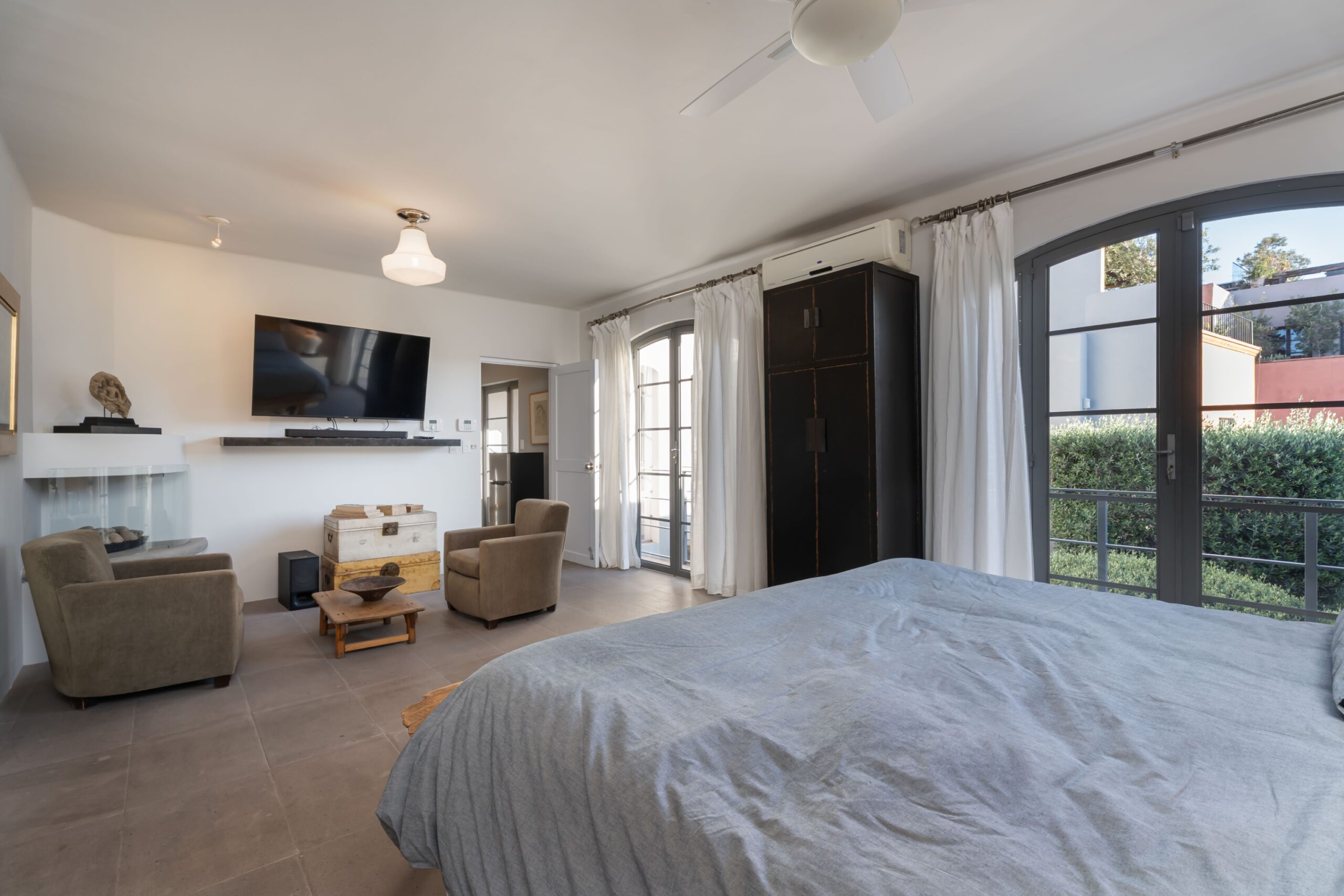 Bright, modern living area with a wall-mounted TV, two beige armchairs, and a bed in a sunlit room with large arched windows and balcony doors, San Miguel de Allende