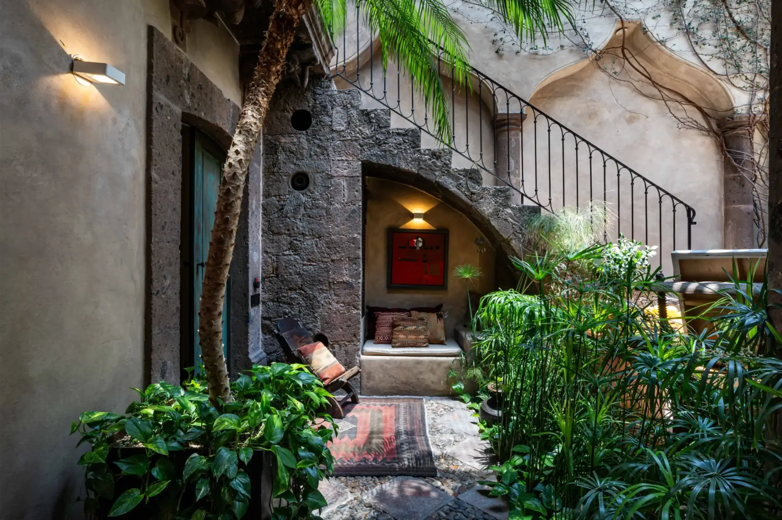 A rustic courtyard with stone walls, a curved iron staircase, and lush green plants surrounding a sitting area with cushions and a patterned rug, San Miguel de Allende