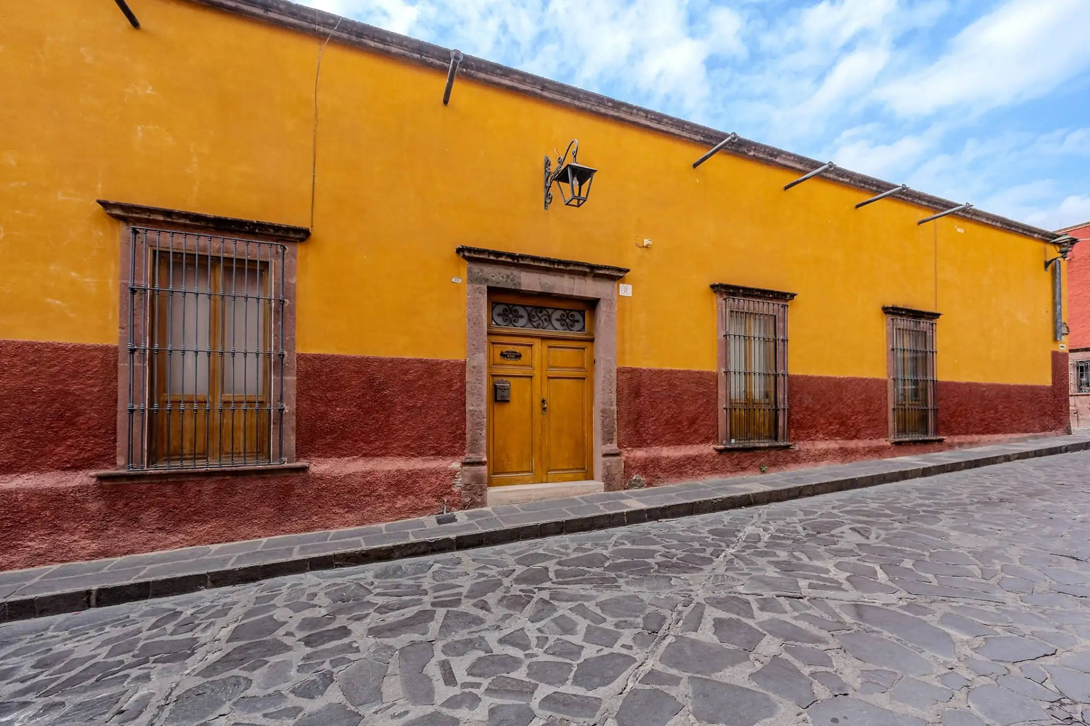 Sunny yellow building with a red base, wooden door, and barred windows along a cobblestone street under a blue sky, San Miguel de Allende